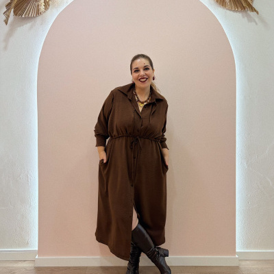 Mulher com vestido comprido castanho e botas pretas de joelho posando frente a parede cor de rosa com arco e decoração seca