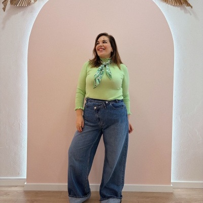 Mulher com camisola verde, lenço no pescoço, calças de ganga largas, em arco branco decorado.