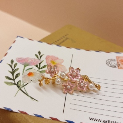 Presilha dourada com flores rosa e pérolas sobre cartão postal com flores desenhadas.