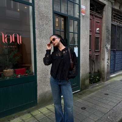 Mulher com blusa preta rendada e jeans azuis numa rua com porta e janela verdes.