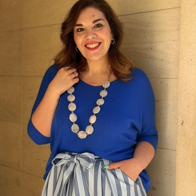 Mulher com suéter azul e saia plissada às riscas junto a parede de pedra bege