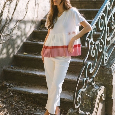 Mulher com blusa branca e rosa plissada, calças brancas, sapatos castanhos, em escadaria de pedra com grade azulada.