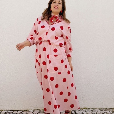 Mulher com vestido rosa claro de bolinhas vermelhas, lenço vermelho no pescoço, sandálias vermelhas, parede branca e calçada portuguesa.