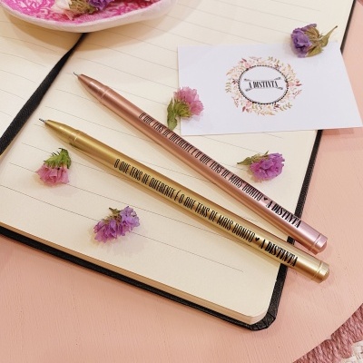 Dois esferográficos metálicos dourado e rosa sobre caderno aberto com flores secas e cartão JANTIVAL