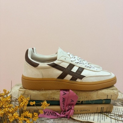 Tênis bege com detalhes castanhos e sola cor de caramelo sobre livros com fita rosa e flores secas amarelas