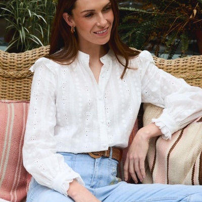 Blusa branca de algodão bordada com botões, jeans azuis, cinto castanho, cadeira de vime com almofadas às riscas