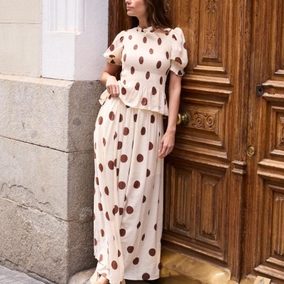 Conjunto creme com bolinhas castanhas, blusa e saia comprida, mulher encostada a porta de madeira.