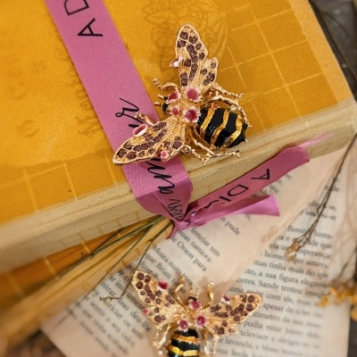 Broches em forma de abelhas douradas com detalhes rosa e preto sobre livro amarelo com fita rosa