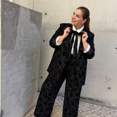 Mulher com fato floral preto e camisa branca de gola rendada num espaço com paredes e chão de pedra.