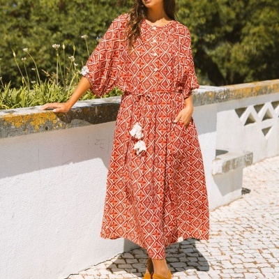 Vestido comprido vermelho e branco com padrão geométrico e cinto com pompons, usado por mulher ao ar livre.