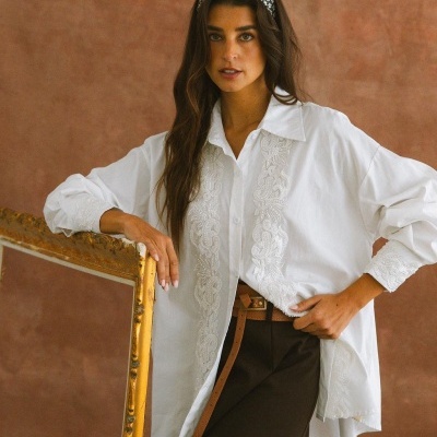 Mulher com camisa branca bordada, cinta castanha e saia preta, apoiada numa moldura dourada com fundo castanho.