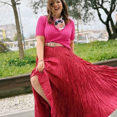 Mulher com saia longa plissada vermelha, camisola rosa e lenço colorido no pescoço ao ar livre