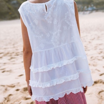 Blusa branca bordada com folhos e saia xadrez num cenário de praia.