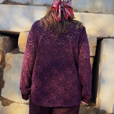 Pessoa de costas com camisola de renda vinho e lenço colorido na cabeça, ao ar livre, com blocos de pedra ao fundo