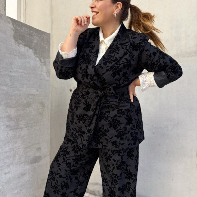 Mulher a usar fato preto com padrão floral em veludo e camisa branca, em ambiente urbano com chão de calçada e parede cinzenta.