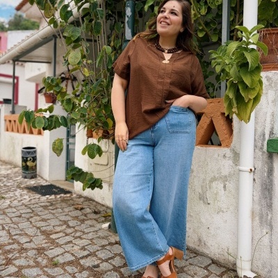 Mulher com blusa castanha, calça jeans azul larga e sandálias castanhas ao ar livre com plantas e paredes brancas.