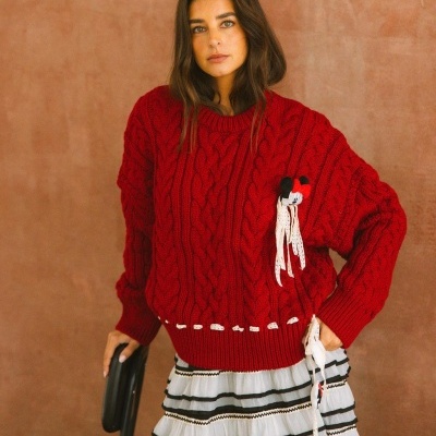 Camisola vermelha de malha com padrão de trança e laços brancos, com saia riscada em fundo bege.