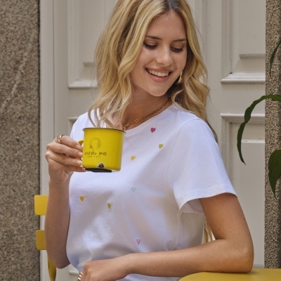 Mulher loira sentada a segurar uma caneca amarela usando uma camisola branca com pequenos corações bordados e jeans azul