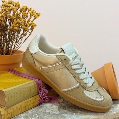 Sapatilha bege com sola castanha e detalhes em camurça em cima de livros amarelos ao lado de planta seca em vaso de barro