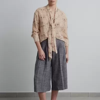 Pessoa a vestir camisa bege floral, calças culotte cinza e sapatos baixos bege em fundo neutro.