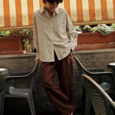 Pessoa com camisa às riscas brancas e cinzentas e calças castanhas em ambiente exterior com cadeiras de plástico.