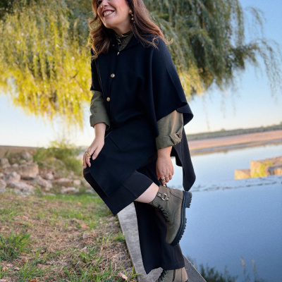 Mulher com capa preta e botas verdes sentada ao ar livre perto de um lago