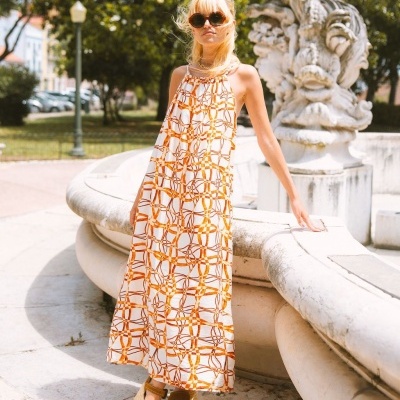 Mulher usando vestido longo branco com padrão laranja, sandálias camel e óculos escuros, ao lado de fonte de pedra