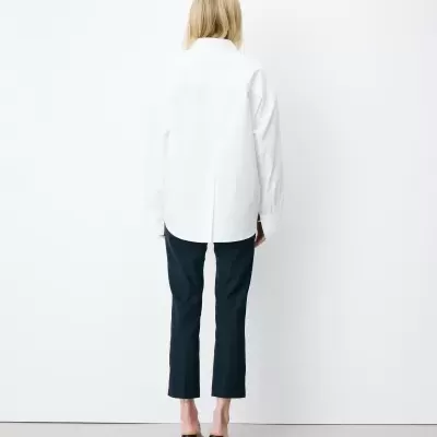 Mulher vista de costas com camisa branca, calças pretas e saltos pretos em fundo branco