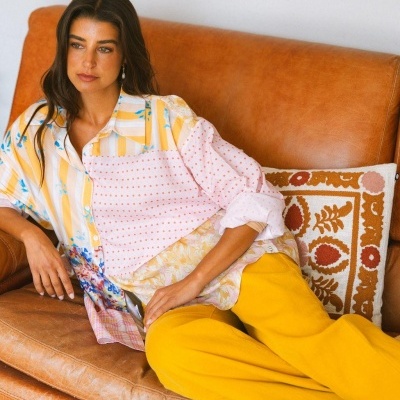 Mulher sentada num sofá de pele castanha com roupa colorida e calças amarelas.