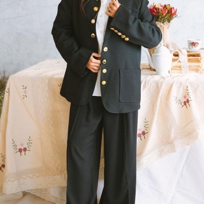 Conjunto de fato preto com botões dourados e camisa branca, à frente de mesa decorada com toalha de renda e flores