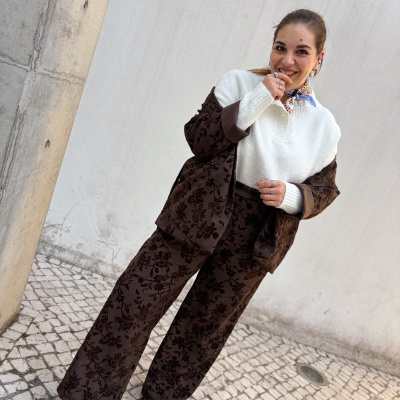 Mulher com conjunto castanho escuro estampado, camisola branca e lenço ao pescoço, posando num cenário urbano com parede branca e calçada portuguesa.