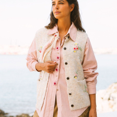 Colete bege com bordados florais usado sobre camisa rosa e calças castanhas em ambiente à beira-mar.