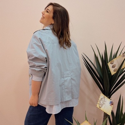 Mulher com casaco cinza claro, camisa às riscas e calças de ganga junto a planta decorativa com cartões postais