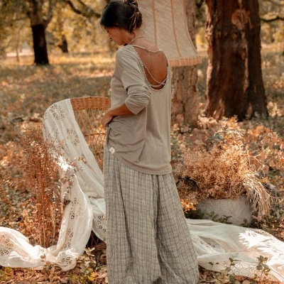 Mulher de costas com camisa bege de decote em V com pérolas e calças largas cinza às riscas em ambiente exterior com cadeiras de vime e tecidos bordados.