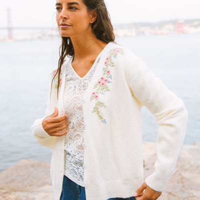 Mulher com casaco branco bordado floral e blusa branca rendada à beira-mar