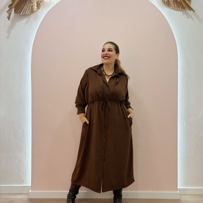 Mulher com vestido castanho comprido e botas castanhas em estúdio com decoração natural no topo e fundo rosa
