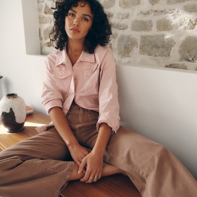 Mulher com camisa rosa e calças castanhas sentada em cómoda de madeira