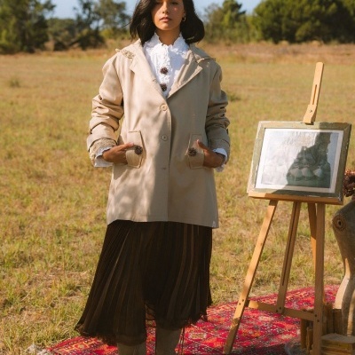 Mulher veste casaco bege com botões decorativos e detalhes em renda, saia preta plissada e botas bege, em campo com tapete vermelho e cavalete de madeira