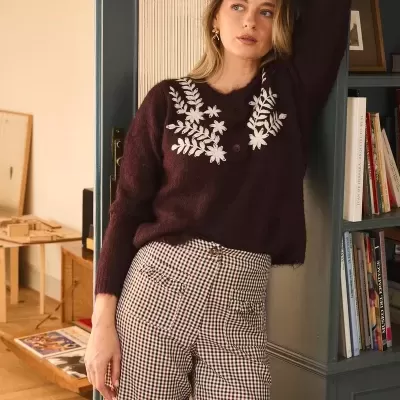 Mulher com camisola castanha bordada e calças xadrez num ambiente interior com estante de livros