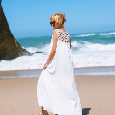 Mulher de vestido branco com croché nas costas na praia, mar e rocha ao fundo.