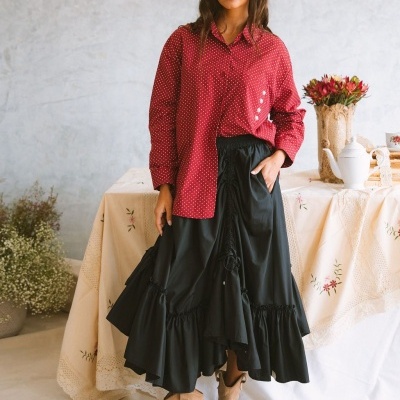 Mulher de camisa vermelha com pontos brancos, saia preta longa e botas castanhas, em ambiente interior com mesa decorada.