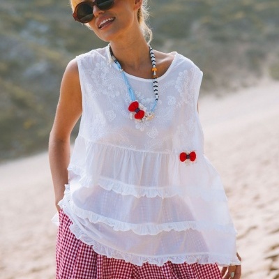 Mulher usando blusa branca folhada, calças xadrez vermelhas e colar colorido, ao ar livre.