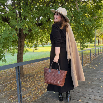 Mulher com casaco bege, vestido preto, botas pretas, chapéu bege e mala castanha trançada outdoors