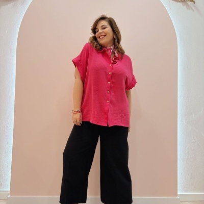 Pessoa com camisa rosa, calças pretas largas e botins castanhos em frente a parede rosa claro com arco decorativo.