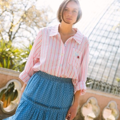 Mulher com blusa riscada rosa e branca com gola e renda, saia azul de bolinhas brancas ao ar livre.
