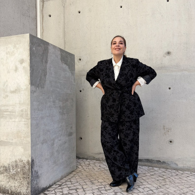 Mulher vestindo um fato feminino preto com padrão floral em veludo, camisa branca de renda e botas pretas, em fundo de parede de cimento e chão com mosaico pedra calcária.