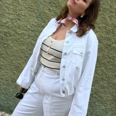 Mulher com casaco branco, calças brancas, top branco riscado, lenço rosa no pescoço e brincos com pérolas.