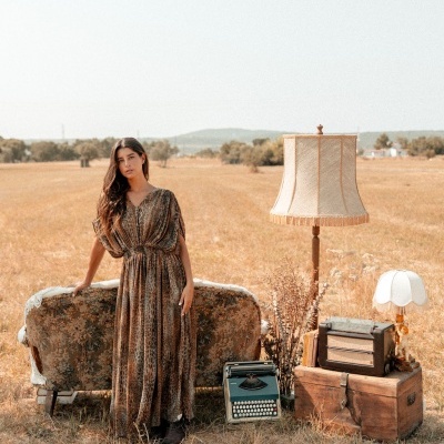 Mulher com vestido longo estampado ao lado de sofá antigo e objetos vintage em campo aberto
