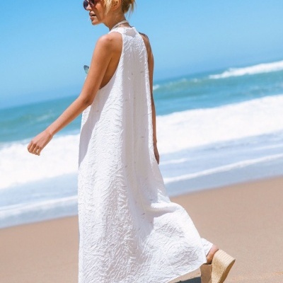 Vestido longo branco com textura e calçado de cunha numa praia com mar ao fundo