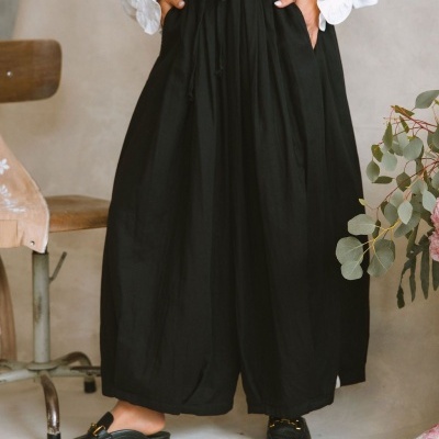 Calças largas pretas com cintura elástica e cordão, combinadas com mocassins pretos e ferragens douradas num ambiente interior.